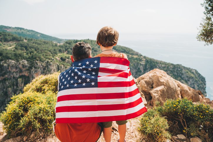 Young man dad father and his son school boy kid standing on a rock cliff with US flag on shoulders and looking at sea. Travellers child wearing American flag on back. 4 July Independence Day.